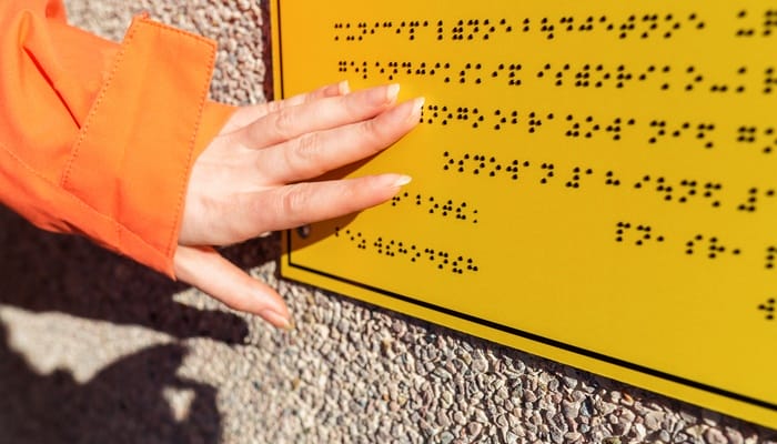 An individual in an orange jacket using their fingers to read the raised dots on a yellow braille sign mounted to a wall.