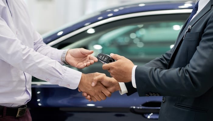A close-up of two suited men's hands shaking as one hands over keys to a new car, signifying a purchase.