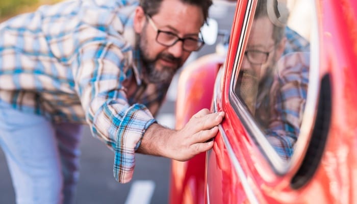 A close-up showing a bearded man wearing glasses and a plaid shirt closely inspecting the window of a red car.