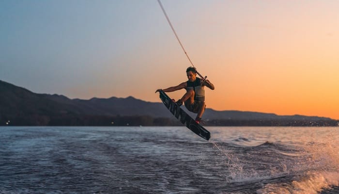 A wakeboarder mid-air after performing a jump. Behind him are mountains lining the lake and an orange sunset.