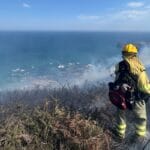 A wildland firefighter wearing protective equipment and a backpack stands among burnt brush overlooking an ocean.