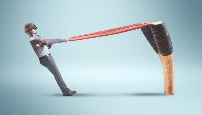 A professionally dressed man in a vintage leather pilot helmet, pulling himself back against the band of a slingshot.