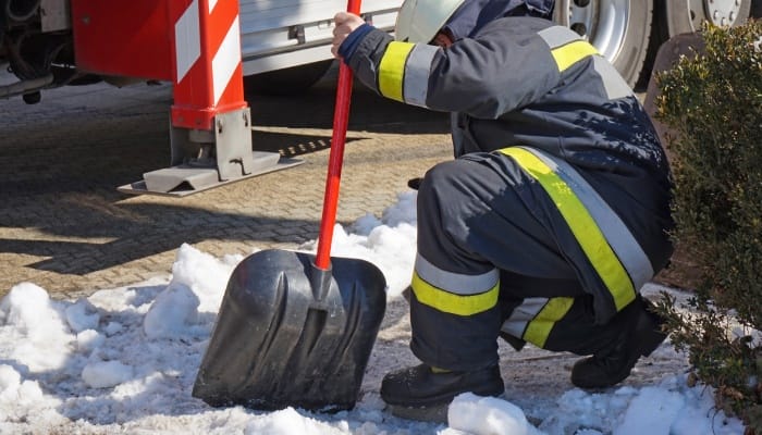 A firefighter wearing a protective suit with reflective stripes and a white hard hat crouches down in the snow, holding a shovel.