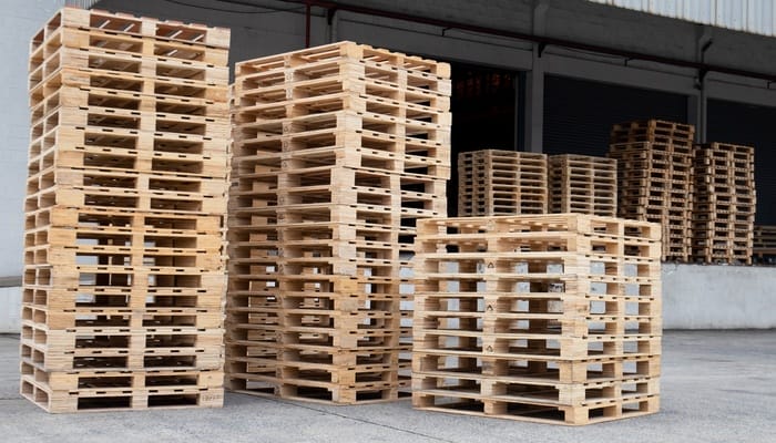 Several tall stacks of wooden pallets sit on a loading dock of a storage warehouse. The warehouse is gray.