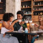 Two men and two women sitting at a table in a cafe with wood shelving. They are holding white mugs in the air.