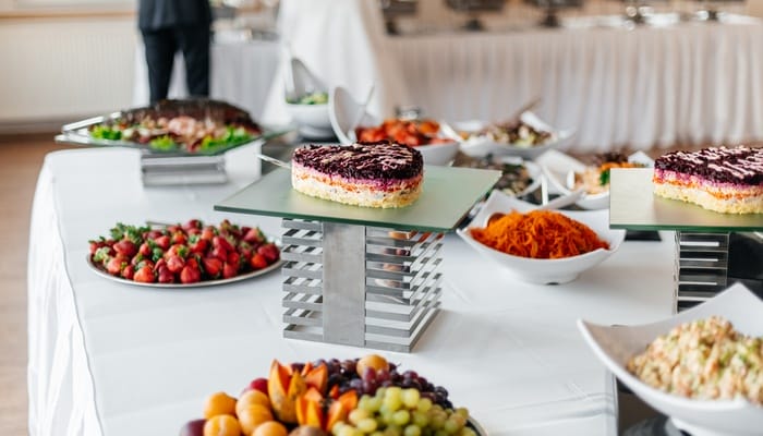 A large table with a white linen covering the top. Food sits on top in separate dishes, including fruit, dessert, and salads.