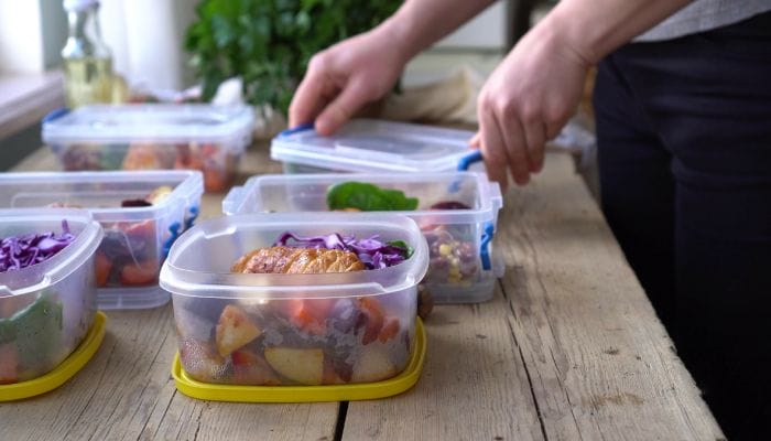 Six meal prep containers filled with food sit on a wooden table. Someone places a lid over one of the containers.