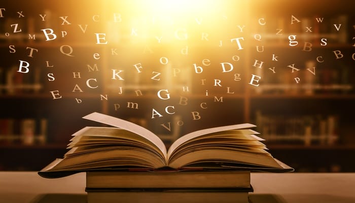 A dictionary sits open on a table in a library. Letters float out of the book and disappear into an orb of light.