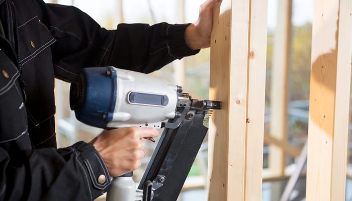 A person wearing a black jacket with white stitching uses a nail gun to attach two skinny wooden beams together.
