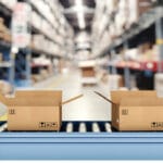 Four small, brown cardboard boxes are going down a conveyor belt in a warehouse. The warehouse is blurry behind the belt.