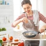 A smiling woman wearing a gray apron and white-and-red striped shirt sprinkles salt into a hot pan.
