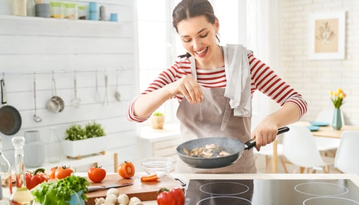 A smiling woman wearing a gray apron and white-and-red striped shirt sprinkles salt into a hot pan.