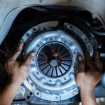 A close-up of a person's hands installing a new clutch plate and flywheel onto a car elevated on a lift.