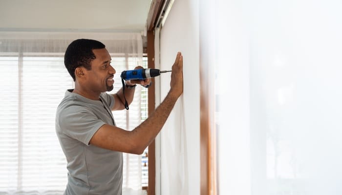 A happy man smiles as he drills the wall for a home improvement project. He's using his right hand to support the wall.