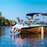 A father and daughter sit on the bow of a yellow and black motorboat while the mother stands behind the wheel.