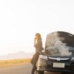 A person holds their phone to their ear while leaning on the side of their car, which has an open hood emitting smoke.