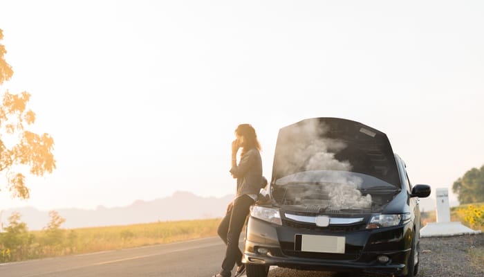A person holds their phone to their ear while leaning on the side of their car, which has an open hood emitting smoke.