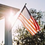 A close-up of a front porch with an American flag displayed on the corner post. The sun shines behind the flag as it waves.
