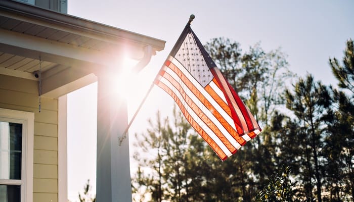 A close-up of a front porch with an American flag displayed on the corner post. The sun shines behind the flag as it waves.