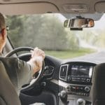 A man wearing sunglasses sits in the driver's seat of a vehicle, steering the wheel as he drives down the road.