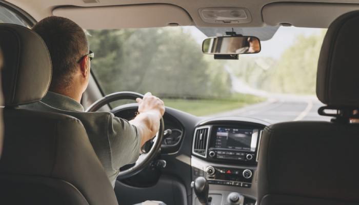 A man wearing sunglasses sits in the driver's seat of a vehicle, steering the wheel as he drives down the road.