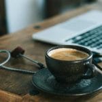A cup of coffee with steam flowing from the top sits next to a laptop and cords on a wooden desk surface.