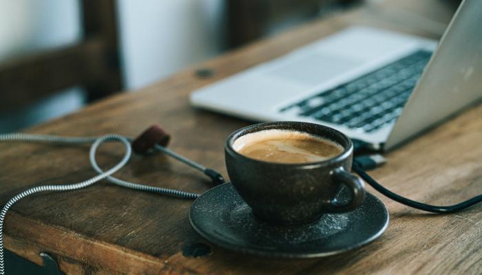 A cup of coffee with steam flowing from the top sits next to a laptop and cords on a wooden desk surface.