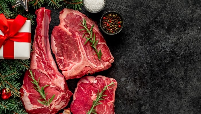 Steak cuts placed beside bowls of seasoning, a wreath with ornaments, and a wrapped gift on a black background.