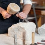 A man wearing an apron using a chisel and hammer to carve into a large piece of white rock on a worktable.