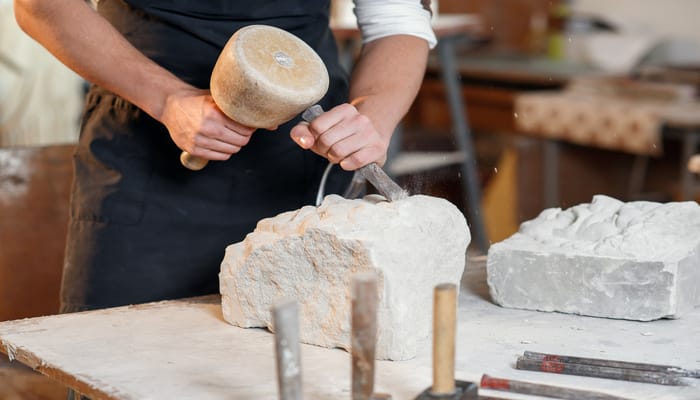 A man wearing an apron using a chisel and hammer to carve into a large piece of white rock on a worktable.