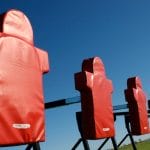 A low view of an American football blocking sled with five red blocking pads on an empty field during the day.