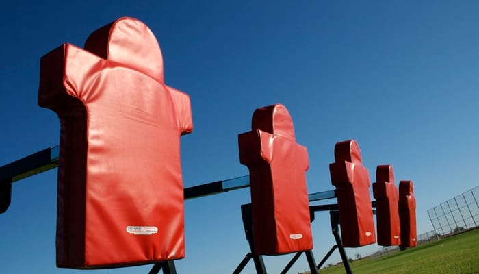 A low view of an American football blocking sled with five red blocking pads on an empty field during the day.