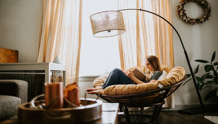 A young woman is reading in a mustard-yellow chair at home, under a large, modern brown floor lamp in her living room.