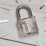 Several lockpicks and a key surround a transparent plastic padlock that sits on a surface of wood panels.