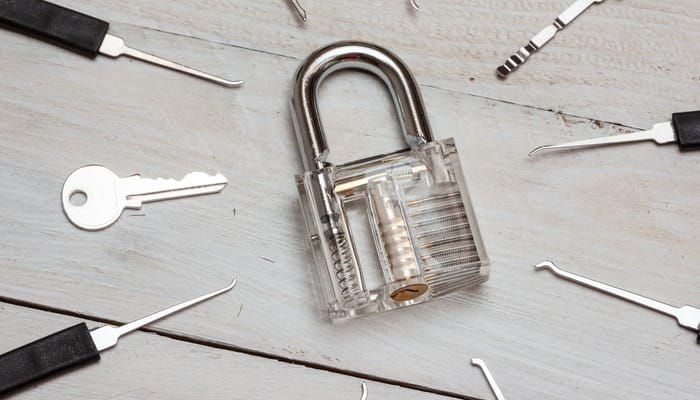 Several lockpicks and a key surround a transparent plastic padlock that sits on a surface of wood panels.