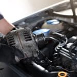 A close-up of a mechanic wearing black gloves and holding an alternator. The car's engine is exposed too.