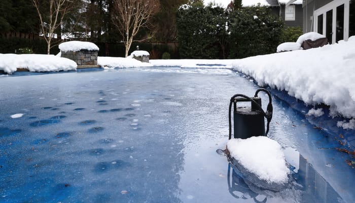 An in-ground pool covered with a layer of frozen water, surrounded by several inches of snow in the winter.