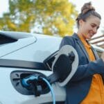 A woman leaning up against her electric vehicle. There is a charging cable going into the car's charging port.