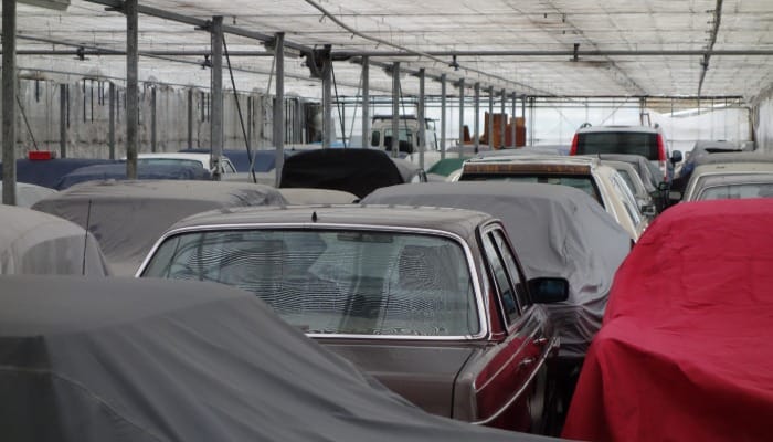 A garage full of stored classic cars. Several of the cars have different colored covers over the top of them.
