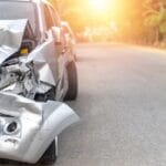 A badly damaged light gray car with its front crushed, parked on the road at sunrise with empty space beside it.