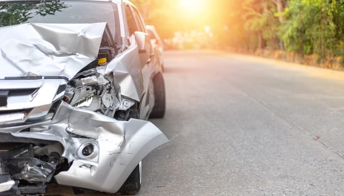 A badly damaged light gray car with its front crushed, parked on the road at sunrise with empty space beside it.