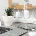 A Laptop and a smartphone are charging on a power strip beside a small potted plant on a home office desk.