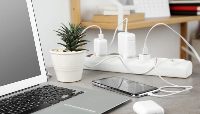 A Laptop and a smartphone are charging on a power strip beside a small potted plant on a home office desk.